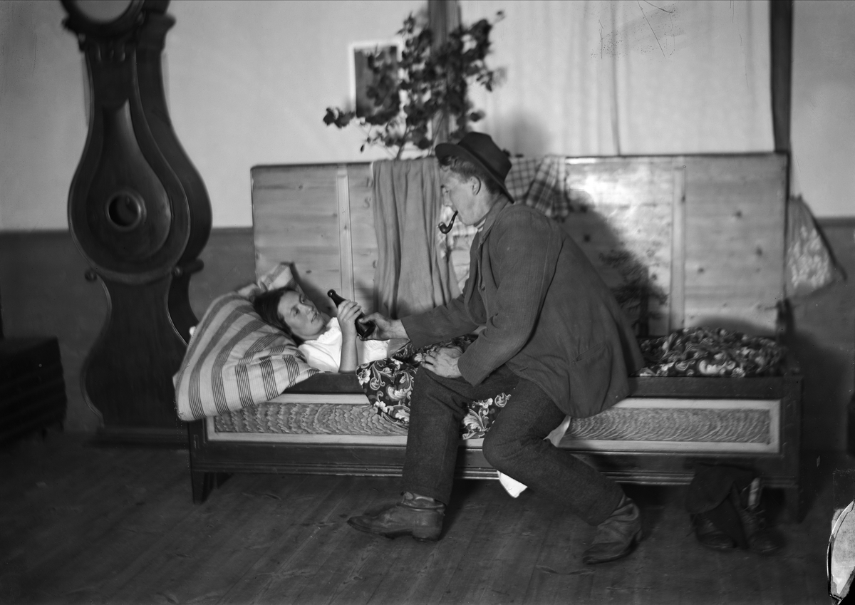 Nattfrieri. En man sitter på sängkanten och pratar med liggande kvinna och bjuder henne en flaska. Värmland, Mangskogs sn.