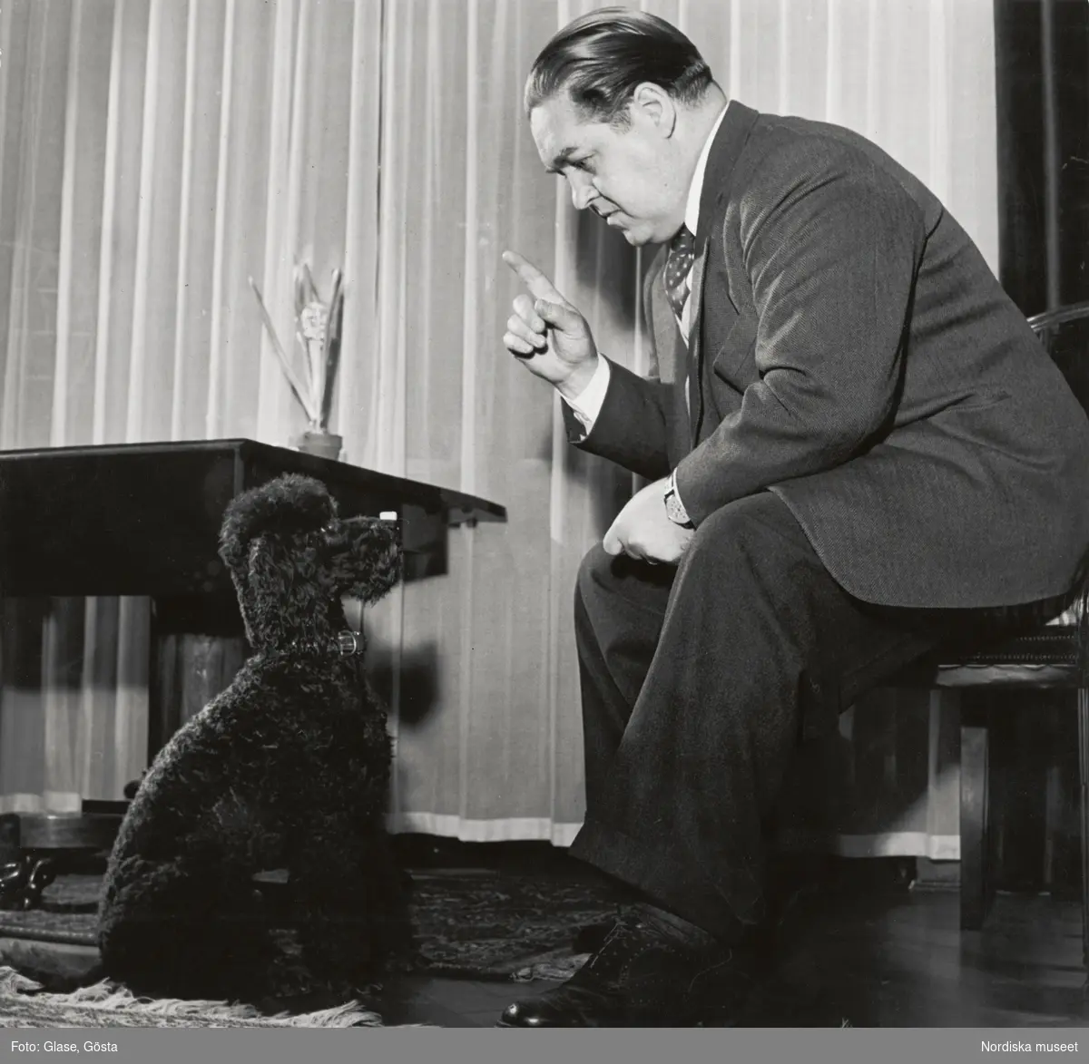 Den svenske operasångaren, tenoren Jussi Björling (1911-1960) avporträtterad tillsammans med en hund. Jussi Björling är klädd i fiskbensmönstrad tweedkavaj, slipover, skjorta och prickig slips. Han sitter i en rumsinteriör och höjer fingret åt en pudel som sitter på golvet framför honom. I bakgrunden ett draperi och ett bord med en hyacint i kruka.