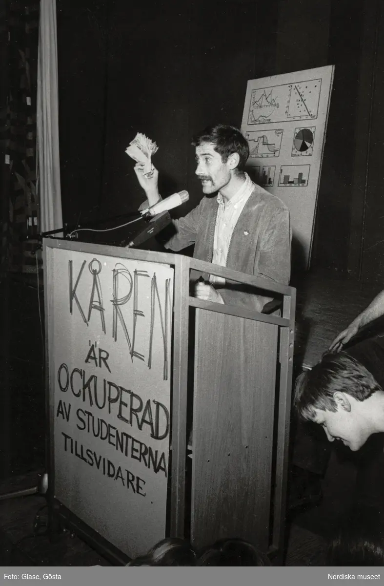Kårhusockupationen vid Stockholms universitet i slutet av maj 1968. I talarstolen på podiet står en ung, mustaschprydd man. Ett plakat bär texten KÅREN ÄR OCKUPERAD AV STUDENTERNA TILLSVIDARE, i bakgrunden en plansch med diagram. Intill står och sitter lyssnande människor.