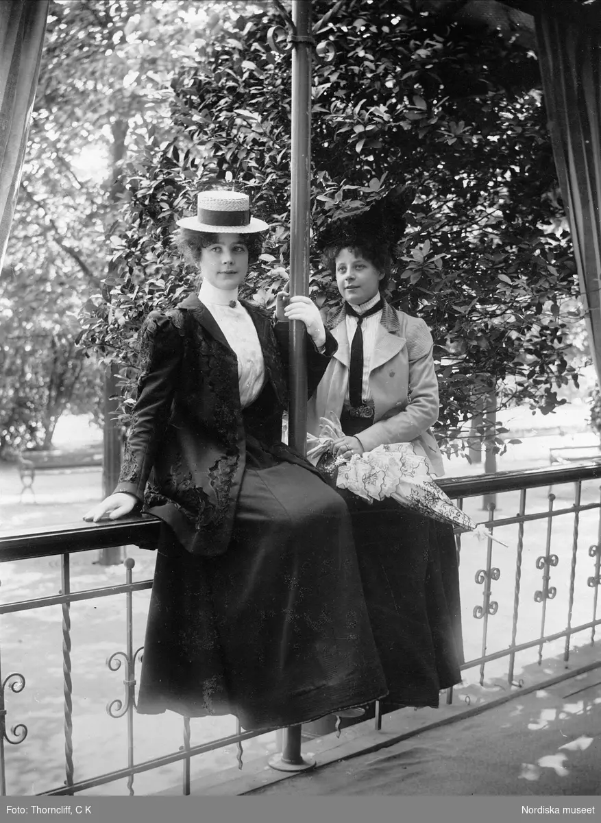 Två unga kvinnor i dräkt och hatt sittande på räcke.