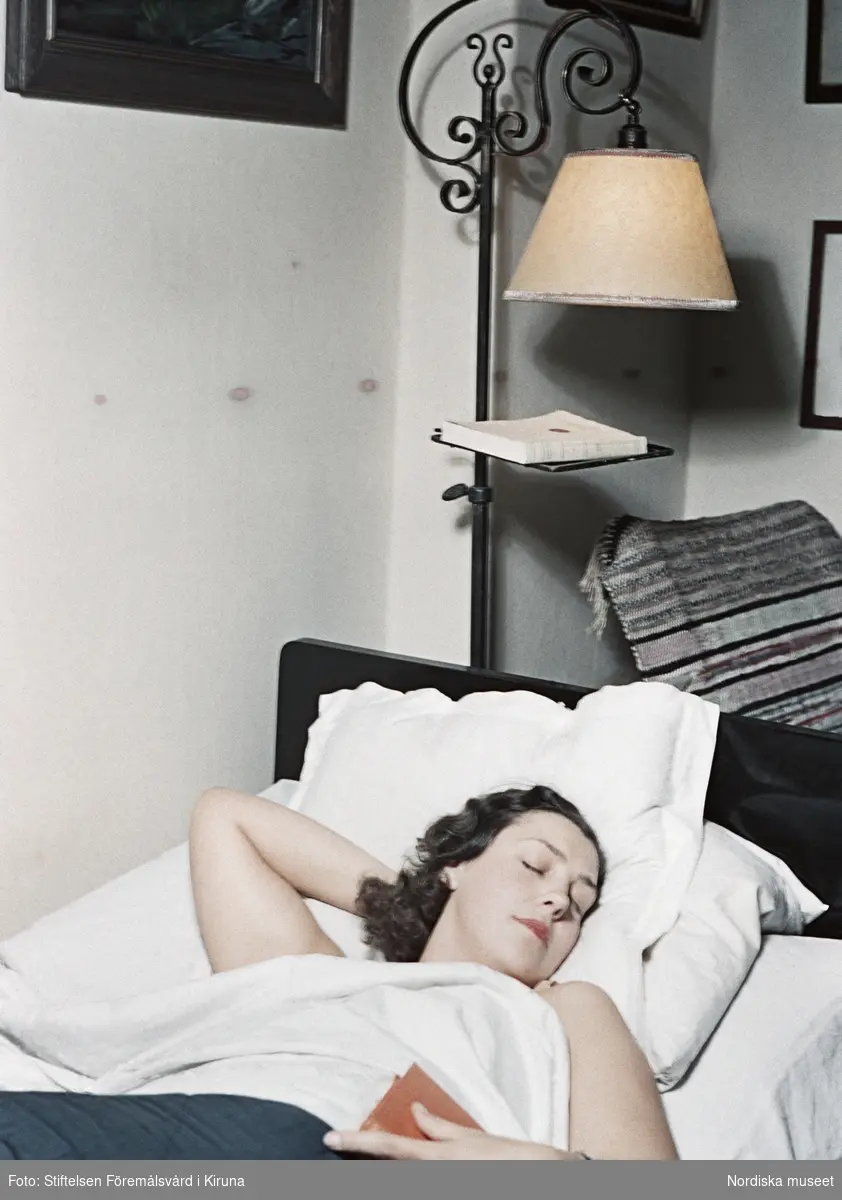 Kvinna med mörkt hår ligger i säng med en bok i handen. Sänglampa.