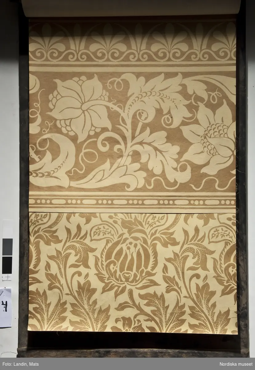 Tapetprov. Skyltstället innehåller 110 tapetprover (25 mönster i olika färgställningar) och 16 tapetbårder (4 bårder i olika färgställningar) på tjockt papper monterade på tunn väv. Varje prov har på baksidan en pappersetikett med tapetens namn och pris samt Libertys firmamärke, lotusblomman, omgiven av texten "LIBERTY:WALL:PAPERS TRADE MARK REGISTRERED". Flera prover utgörs av polykroma blommönster med maskrosor, pioner och löjtnantshjärtan mot ljusa bottnar, men de flesta proverna är storskaliga damastmönster med akantusblad och palmetter, ofta i två nyanser där botten är mörk och motiven ljusa eller tvärt om. 1800-talets andra hälft.