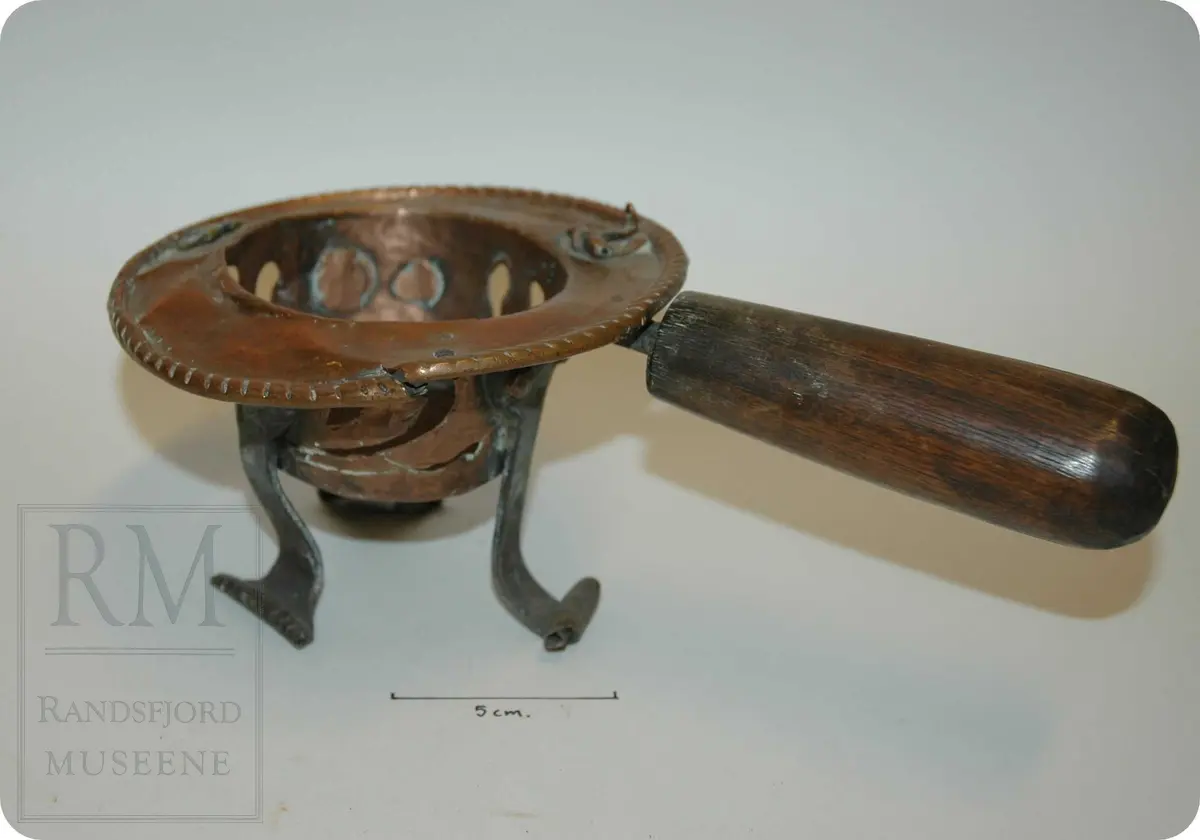 En messing"kopp" med brede kanter, med tre føtter av jern, samt et kraftig trehåndtak. mye dekor på "koppen"/beholderen.