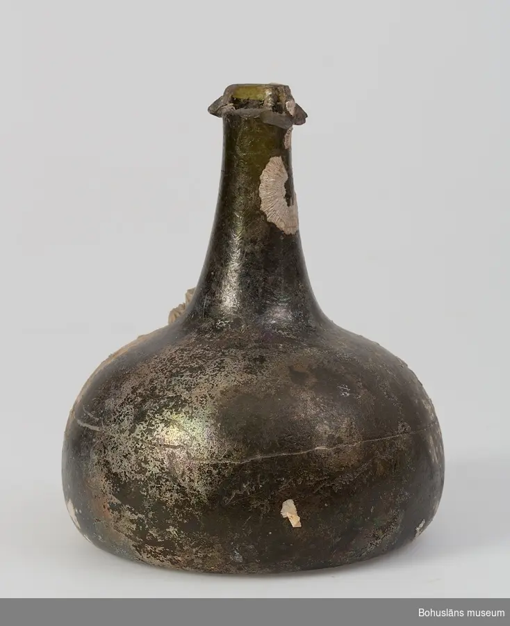 Flaska av mörkgrönt glas, klotformad med konad hals. Påklippt halsring, regalin. Stucken botten (uppåtstående) med rått puntelmärke. Mer än halva  halsringen är avslagen. Flaskan har iriserat glas och en kraftig påväxt av havstulpaner.

Antecknat i Gåvobok för  Uddevalla museum februari 1885:
"-2ne glasflaskor, deraf den ena bär årtalet 1744, [UM000692] den andra legat öfver 100 år i hafvet, då den upptagits från det Svenskt-ostindiska skeppet "Göteborg", hvilket sjönk år 1745. - På denna senare sitta en del snäckor [överstruket och ersatt med 'kräftdjur lepas-art']. -Gifvaren antager att flaskorna brukats för kneif eller soja.-"

Ur handskrivna katalogen 1957-1958:
Sojaflaska
Bottendiam.: 12,5. H. 15,3. Av mörkt glas (med snäckornament?) Skadad, särskilt halsen. (10)