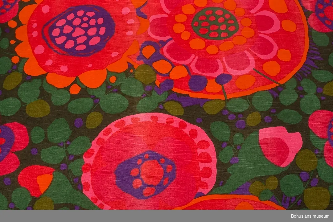 Funktion: Rumsavskiljare i tångbadhus.
Draperi av bomull, tryckt blommigt mönster i grönt, rött, rosa och lila på ripsartat (inslaget)vävt tuskafttyg.
Ljusare slitna ränder, enstaka hål.
Mönstret heter "Ekerö", och ritades av Saini Salonen runt 1969 till kollektionen "Linje Hemma" för Borås Wäfveri.
