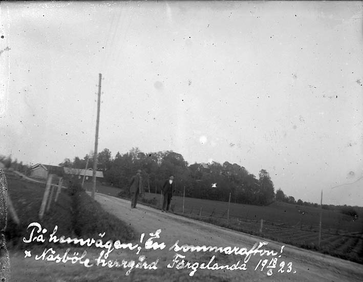 "På hemvägen! En sommarafton! Näsböle herrgård Färgelanda 18/6 1923"
Två män i kostym och hatt går längs en väg genom ett jordbrukslandskap