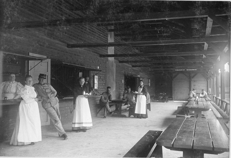 Marketenteri med tre serveringluckor och öppen veranda. Soldater och markenterskor med serveringsbrickor poserar för fotografen. På mittenluckan ligger en hund, Uppsala 1895.