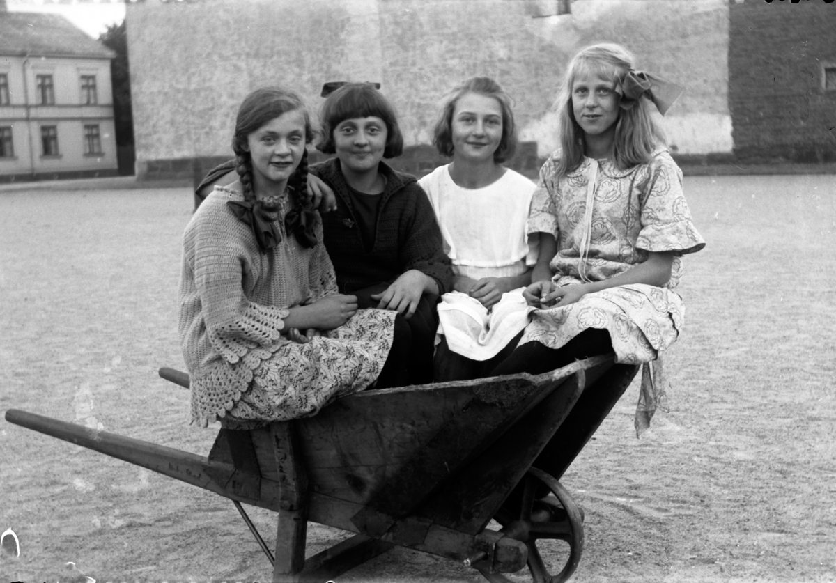 Fyra flickor sitter i en skottkärra på Västra torget i Jönköping. Maj Karlsson (1) född 1916, Majken Karlsson (2) född 1918, Mildred Svensson (3).
