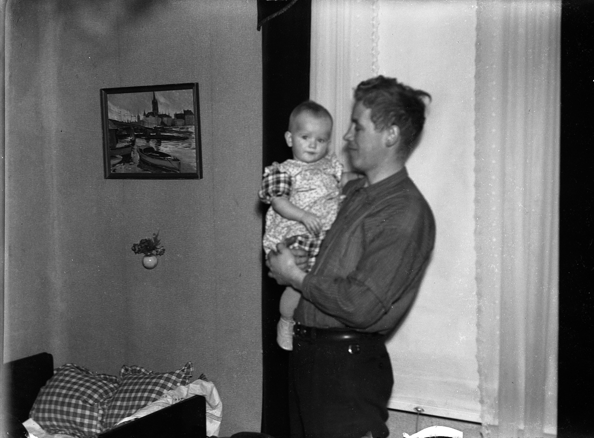 En man med ett litet barn på armen står framför ett fönster med nerdragen rullgardin.