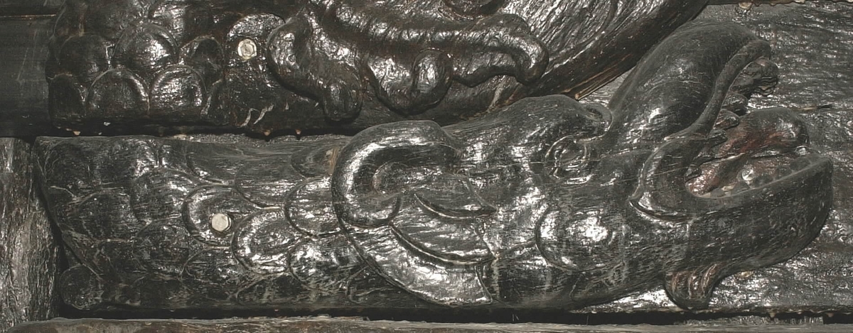 Skulptur i form av en drake (delfin?), återgiven i höger profil.
Draken har ett stort huvud med uppåtvriden nos och vidöppen käft, blottande en tjock tunga och spetsiga tänder. Halvrundade vingar. Stjärten fortsätter uppåt utmed stjärten till en triton, se fyndnummer 00020. Baksidan är slät.
Skulpturen är mycket välbevarad.

Text in English: A sculpture of a dragon or dolphin in right profile.
The dragon has a large head with upturned nose and wide-open mouth, exposing a thick tongue and pointed teeth. Semi-round wings. The tail extends upwards with the tail to a Triton, see No. 00020. The back is smooth.
The sculpture is well preserved.