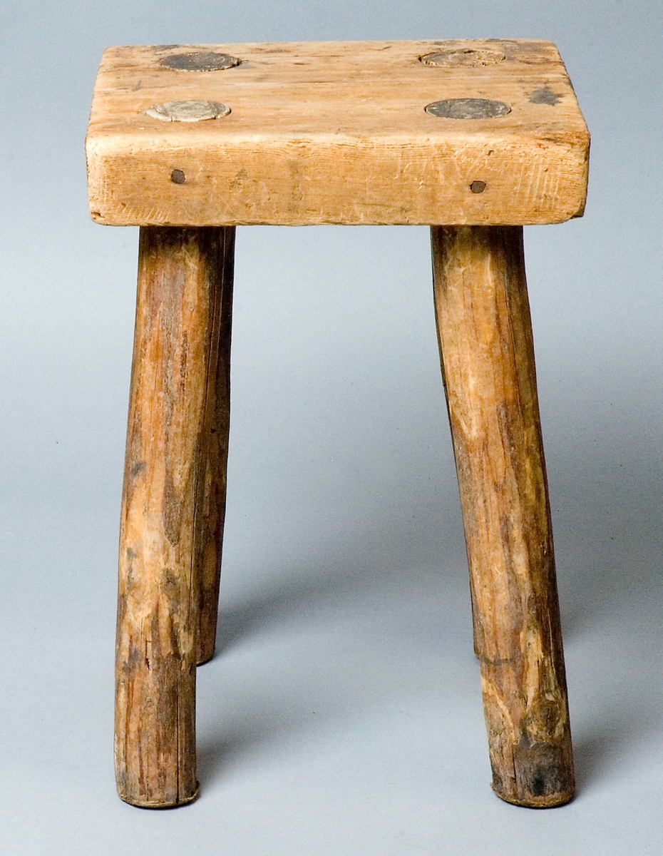 Pall, handgjord av trä, furu. Fyrkantig sits med fyra genomgående itappade ben. 
Skrivet under sitsen: Skansens rekvisita (Delsbogården) BOLLNÄS.