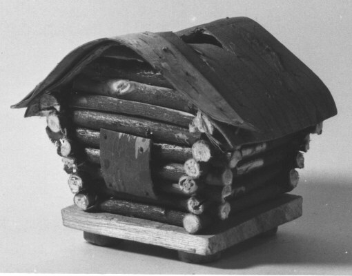 Sparbössa av trä, i form av ett timrat härbre. Taket av
näver, där finns även myntinkastet. Huset står på en träplatta på
fyra fötter. Sparbössan var ett av många bidrag till en tävling
arrangerad av Postbanken 1942. Flickorna skulle göra omslag till
motböcker och pojkarna sparbössor. Bidragen ställdes sedan ut på
Postmuseum. Denna sparbössa är gjord av Folke Dahlgren, Ursviken.