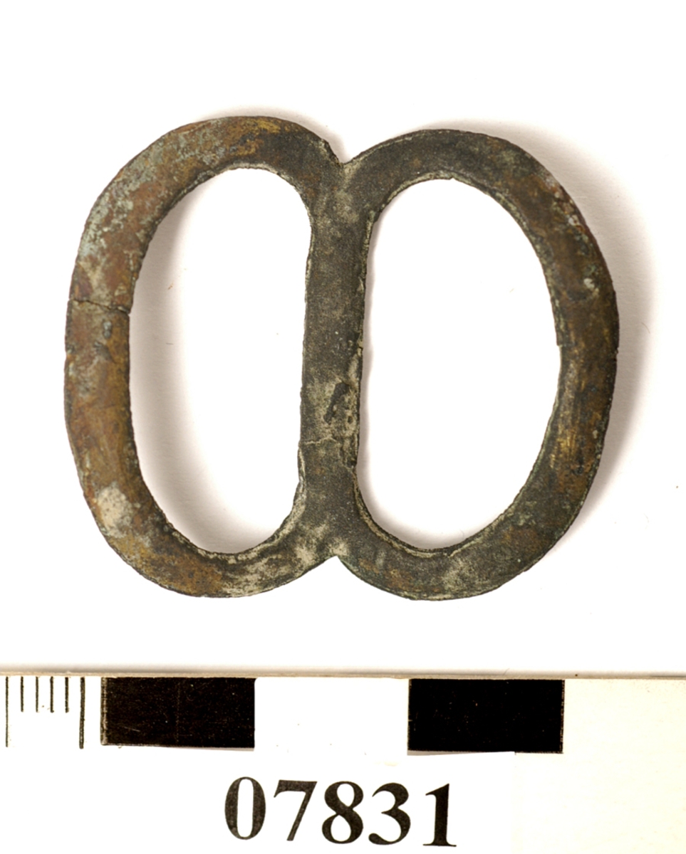 Sölja, spänne. Formad som en åtta. Lätt böjd på mitten.

Text in English: One metal buckle.
One side of metal (probably front) is convex, the other side (probably back) is flat, with defined edges.
The whole piece is concave, the center bar sits lower than the sides. When viewed from the side, it resembles a wide V. When viewed from the front or the back, it resembles a flattened figure 8.
Handmade out of what appears to be one piece of metal, two solder points visible.