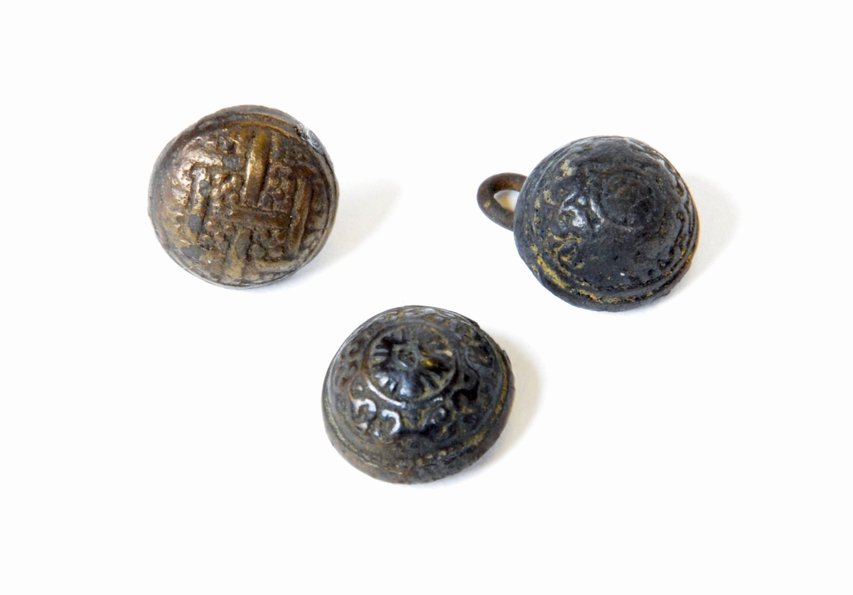 Skoknappar, 3 stycken av mässing.
2 med ögla, 1 utan. Ornering på knapphuvud (varierande).

Text in English: Three decorated metal buttons.  Two with similar but different patterns.  All are half-sphere shape with almost flat bottoms.  Two have wire shanks, one is missing.
First pattern is a lattice.  Six lines are woven together and each space between is filled with five dots. This button has a wire shank that is cast in far offcenter.
The second button has a flower at the top in the center with a circle around it.  Under this is a pattern that is too worn to be accurately described, but appears to be a four-repeat swoop pattern.  The wire shank is cast into the head, and is severely bent to one side.
The third button has a five-petal flower in the center at the top with a circle around it.  The pattern below it is a five-repeat pattern of opposing "C"s, connected by a line in the center and with dots above and below the line.  There is no shank, but holes in the bottom indicate there was probably a wire shank at one time.