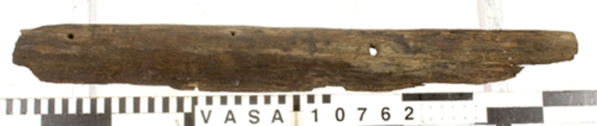 Bräda.
Ena långsidan mycket skadad. Avbrutna ändar. Tre genomgående spikhål.

Text in English: One beveled long edge. One end is tapered, which might have been for a reinforcing iron.
Two iron nail fastenings. One iron bolt or treenail with carved head.
Surface erosion and breakage.