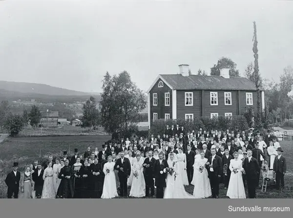 bröllopsgäster och brudpar församlade framför fmangårdsbyggnaden. Majstång i bakgrunden.