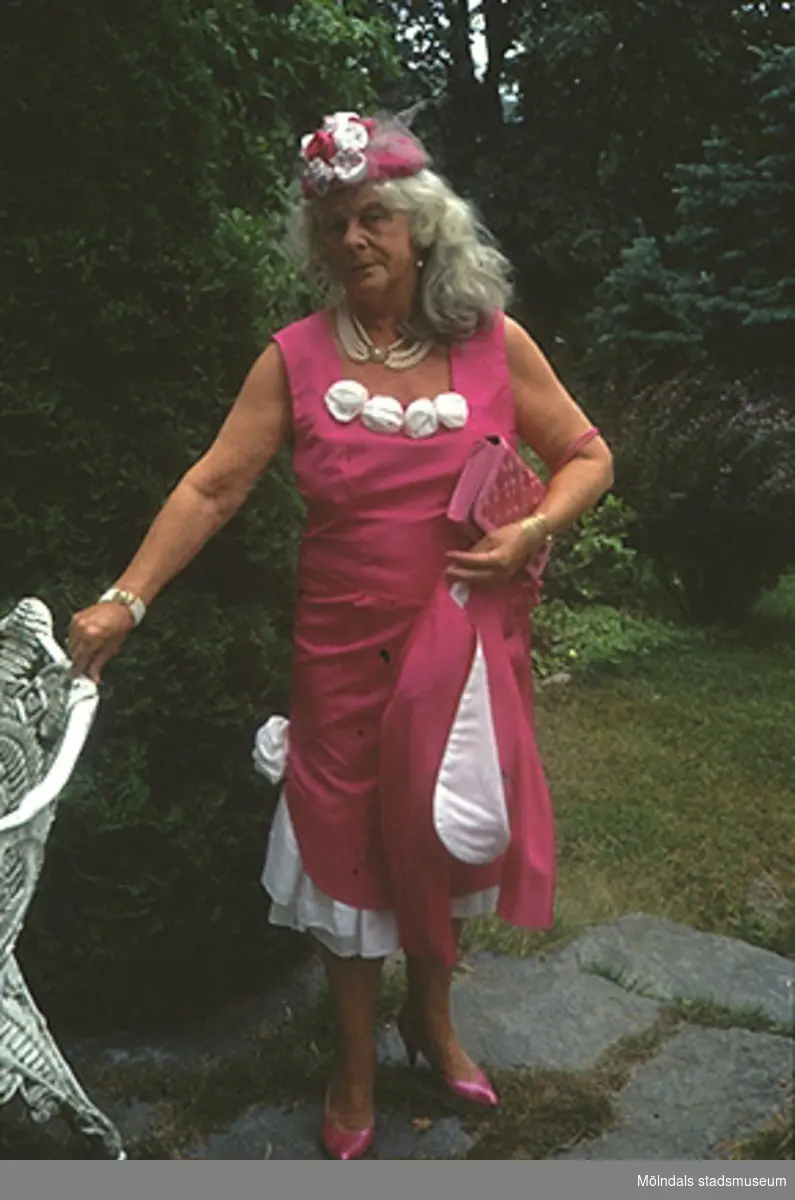 Margit Wannerberg står i en trädgård, iförd sin 1990-tals dress. Plaggen finns på museet: klänning/jacka/sjal (inv.nr: 03335_1-3), handväska (invnr: 03336), hatt (inv.nr: 03339) och skor (inv.nr: 03337).