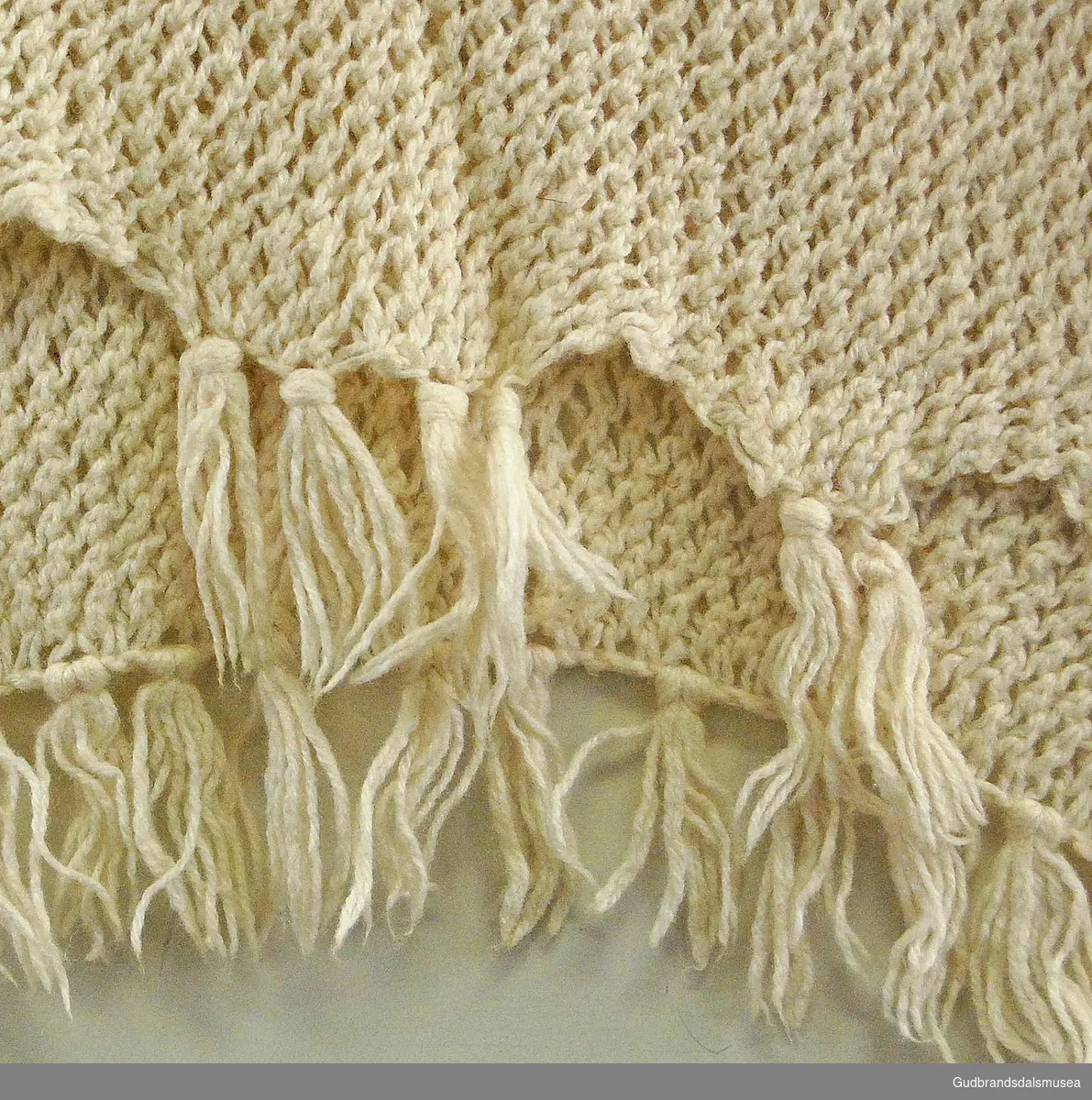 Knyttet/strikket i hvittbeige ull, med knyttede frynser i endene. Bredt skjerf, kan lett utvides. I rutetmønster; skråstilte løkker langs hele skjerfet.