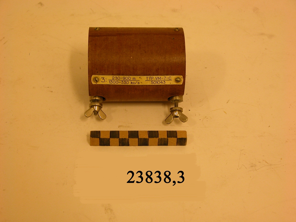 Ljusbrun cylinder med dubbla väggar och två skruvar av metall med vingmuttrar. På cylindern en fastskruvad bricka av plast med text : 3: 230-900 m. 1300-330 kc/s. TYP VM-7-C. 501043.
