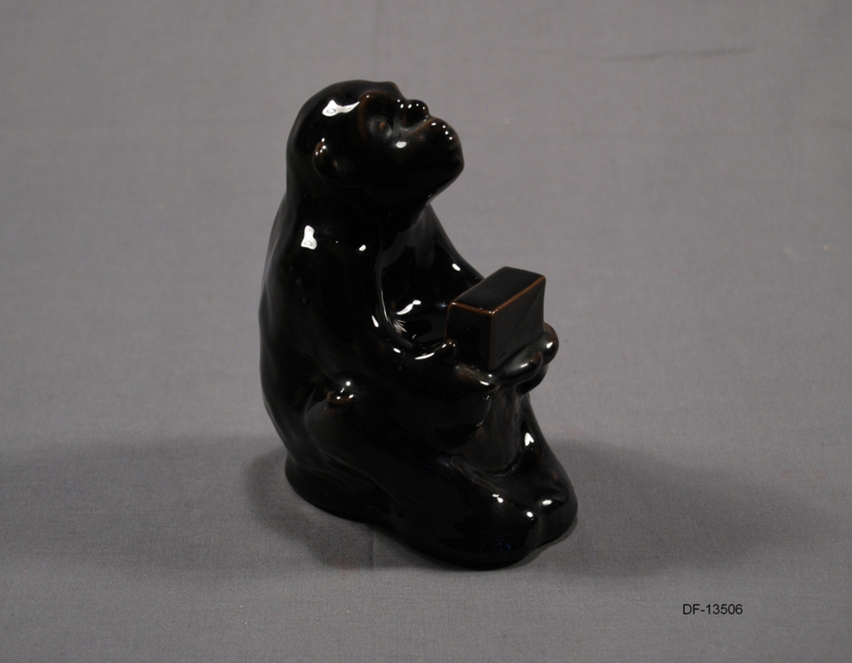Keramikkfigur utformet som sittende ape. Armene er foldet og holder en liten boks. Fungerer som holder for fyrstikkesker.