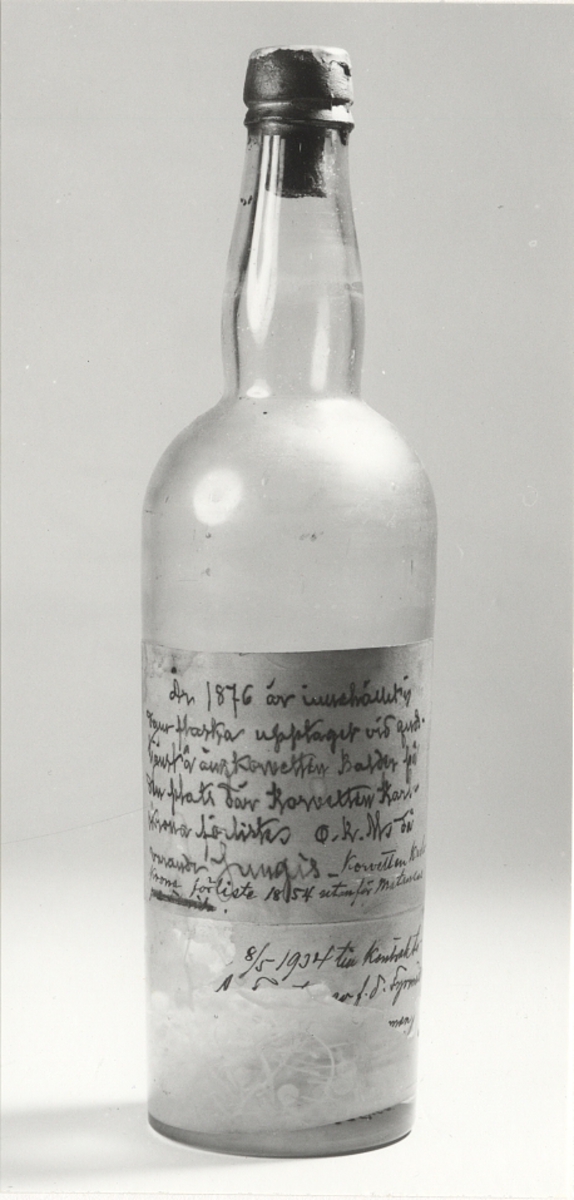 Etikett på flaskan: "År 1876 är innehållet till denna flaska upptaget vid gudstjänst å ångkorvetten BALDER från den plats där korvetten KARLSKRONA förliste. O K Ms dåvarande Jungis. Korvetten KARLSKRONA förliste 1854 utanför Maranzos 8/5 1934 till kontrakts-... av f d Fyrmäst..."