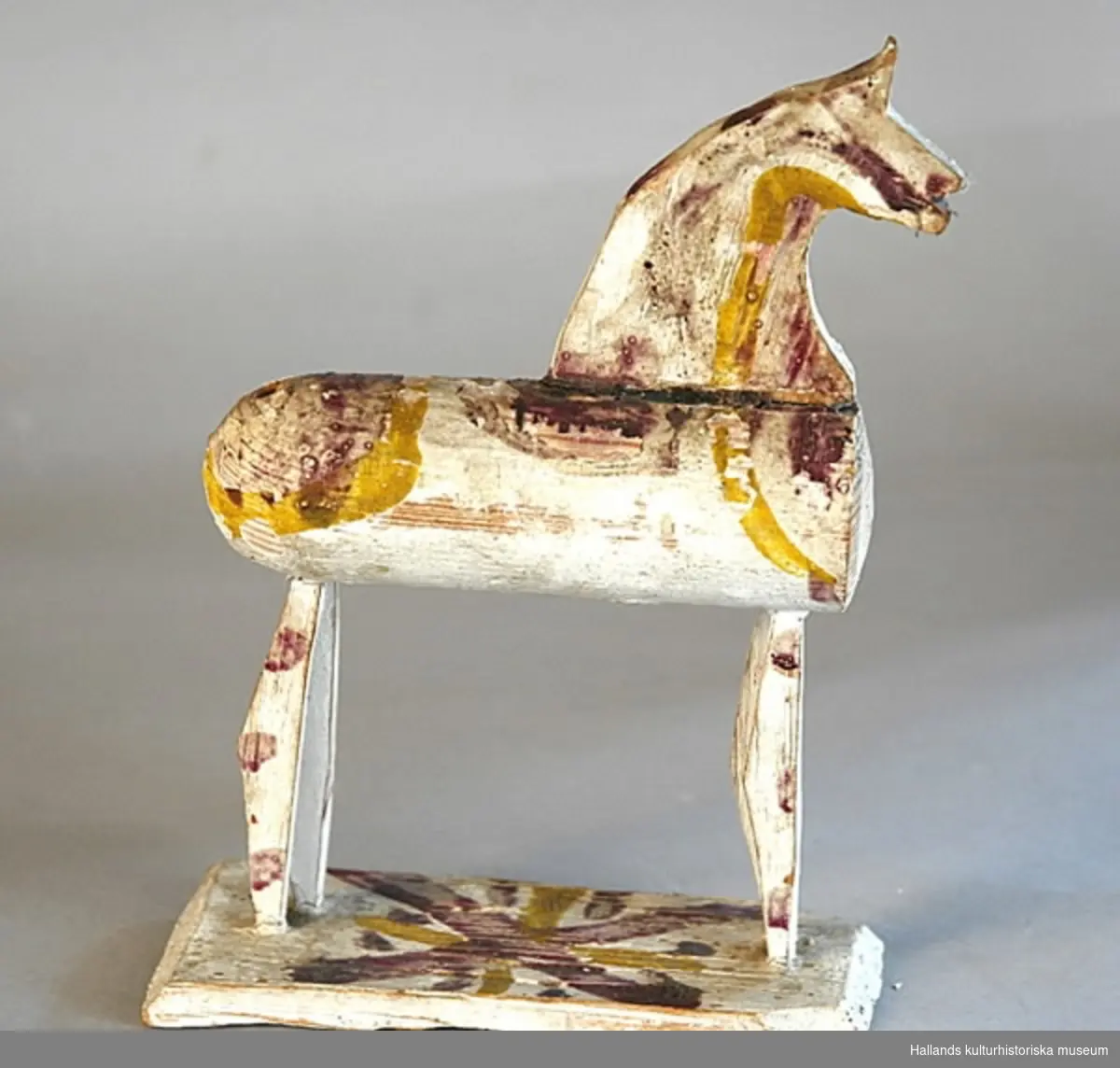 Leksakshäst av trä, vitmålad med mönster i gult och gredelint. Kroppen cylindrisk, svansen har fallit bort. Huvudet en profil utskuren i en träskiva, benen likaså. Hästen står på en bräda.