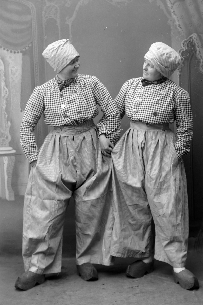 Studioportrett i helfigur av to kvinner, iført kostyme bestående av rutete skjorter, posete bukser, hodeplagg og tresko.