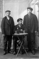 Studioportrett av tre menn samlet rundt et bord med flaske o
