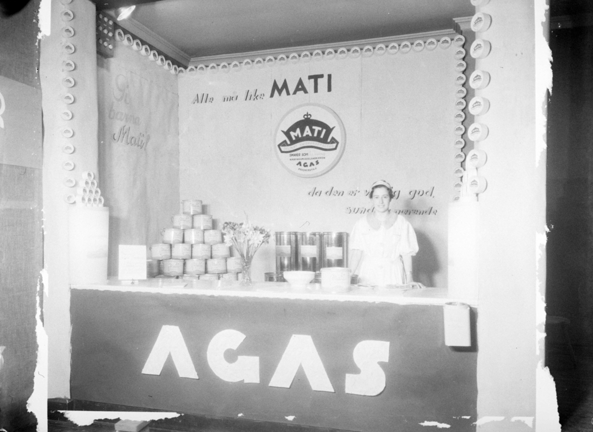 Reklame for produsenten AGAS sitt smørbrødpålegg "Mati". Utstillingsstand med varer og pike som presenterer. Reklametekst: "Alle må like MATI da den er veldig god, sund og nærende" og "Gi barna Mati!"
