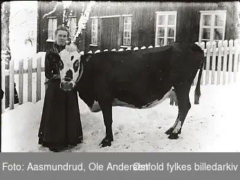 Kvinne med melkeku på framside av våningshuset på gården Aasmundrud. Vinter og snø. Kvinne ukjent.