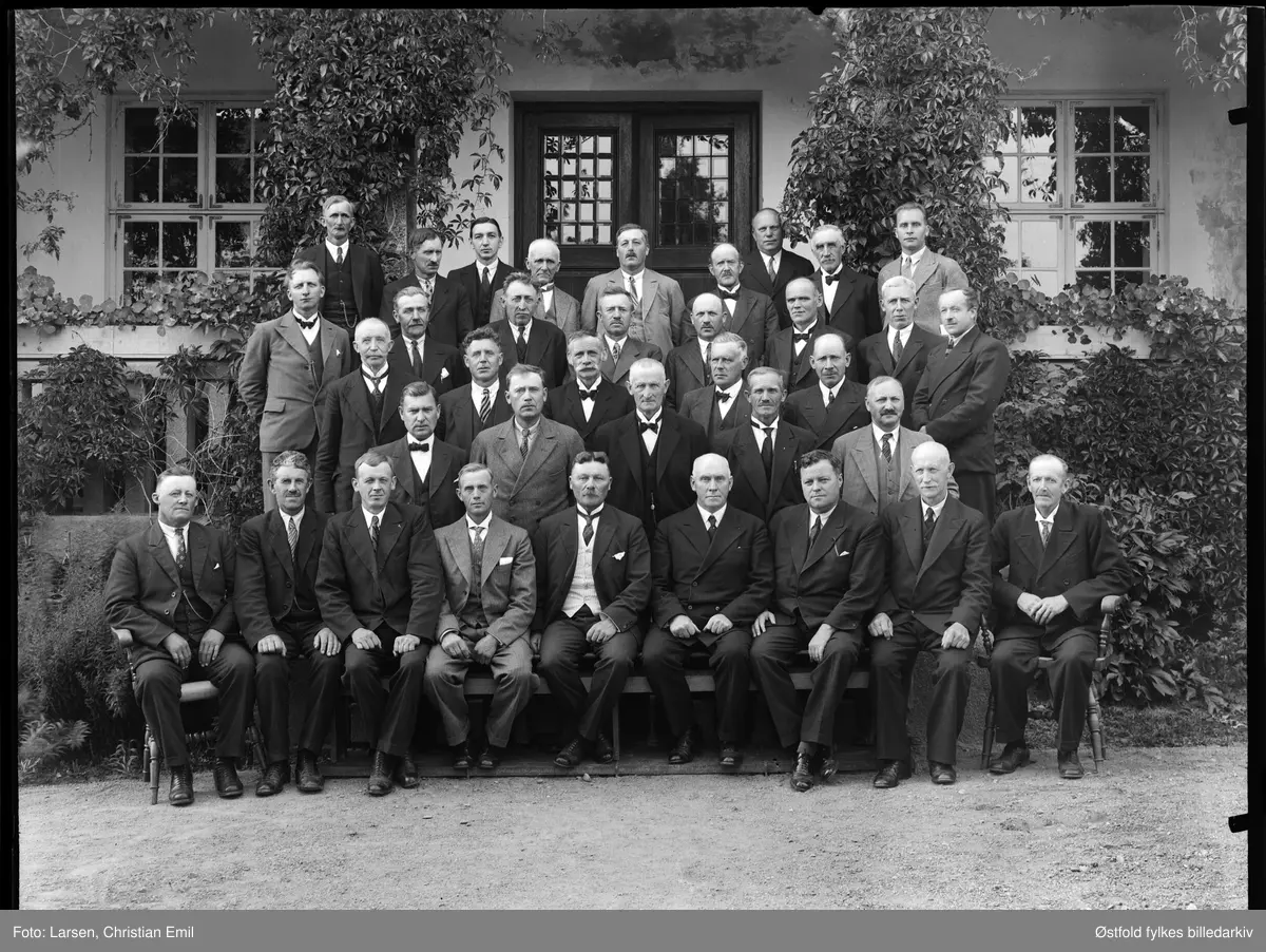 Gruppe menn sittende utenfor et stort hhus, samme motiv på plate i strr. 12x16 cm.
Rakkestad og Degernes