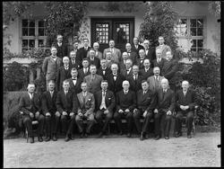 Gruppe menn sittende utenfor et stort hhus, samme motiv på p