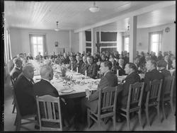 Ukjent gruppe menn rundt dekket festbord, våpenskjold på veg