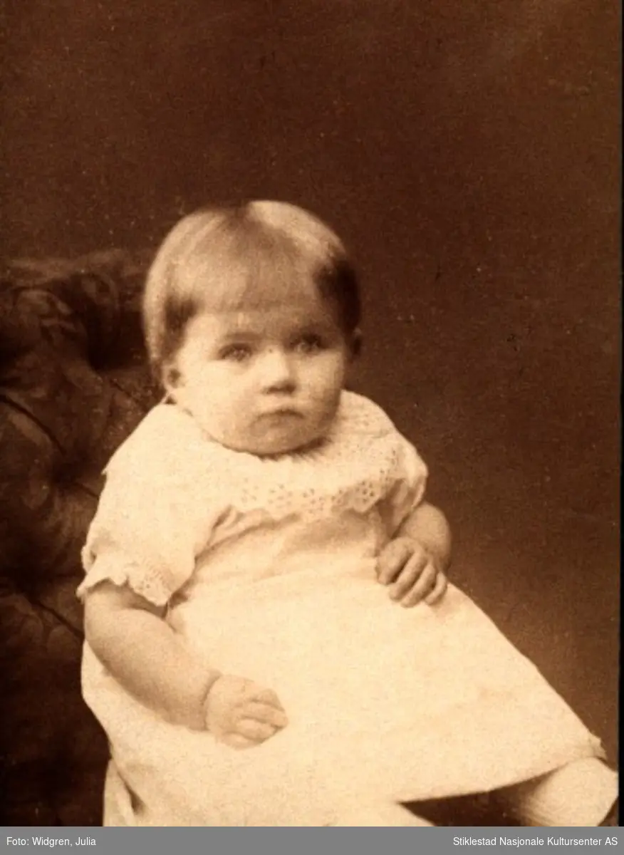 Portrett av Gunhilde Moe som barn i lys kjole med blonde, sittende i stol