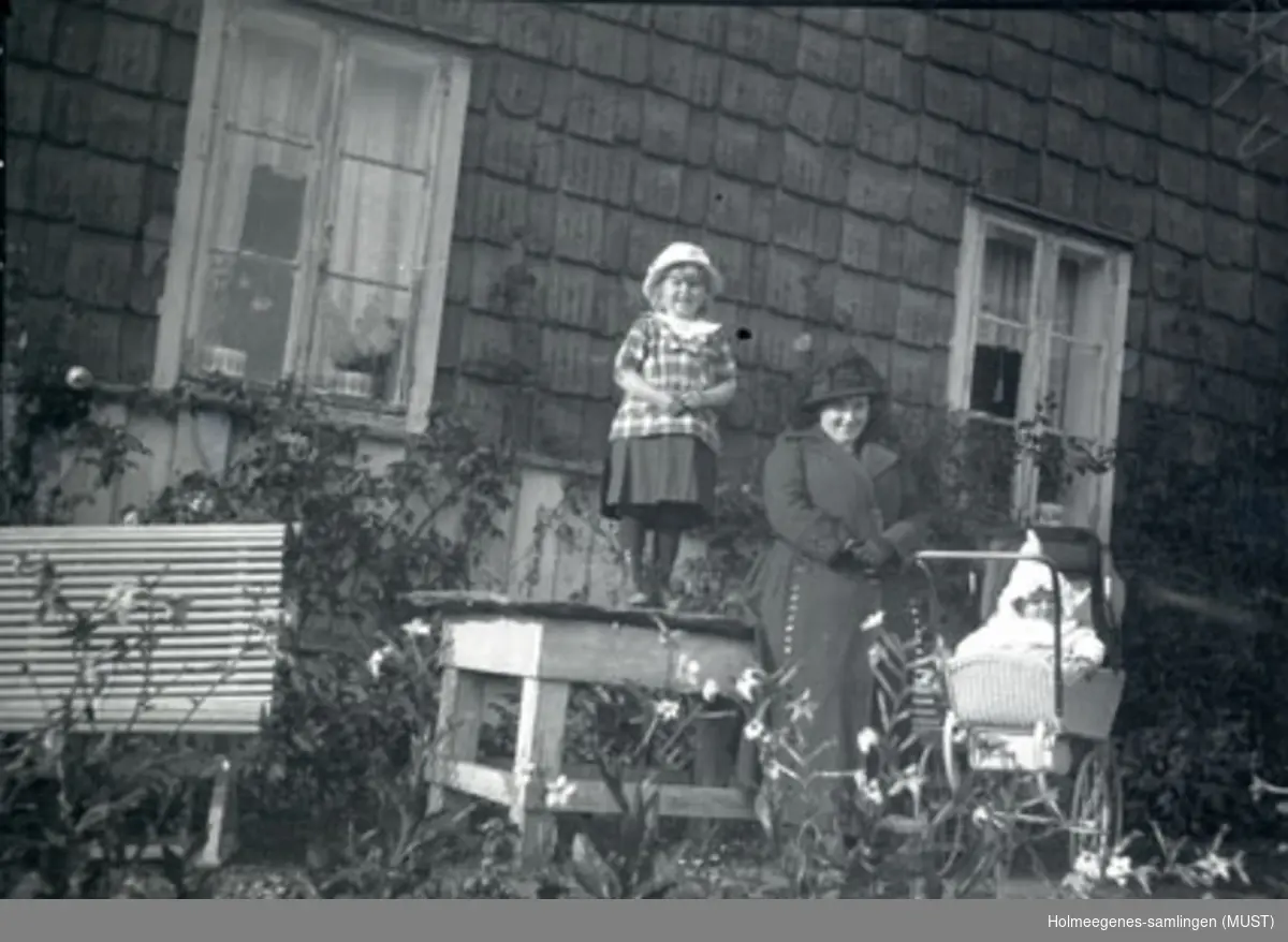 En voksen kvinne med et barn i barnevogn, og en liten jente som står på et hagebord, foran huset på Holmeegenes i Stavanger.