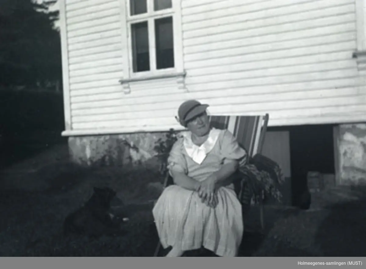 En kvinne i sommerkjole og hatt sitter i en solstol utenfor et bolighus. Antatt samme sted og tidspunkt på ST.K.HE 2010-011-0091 til -0094.