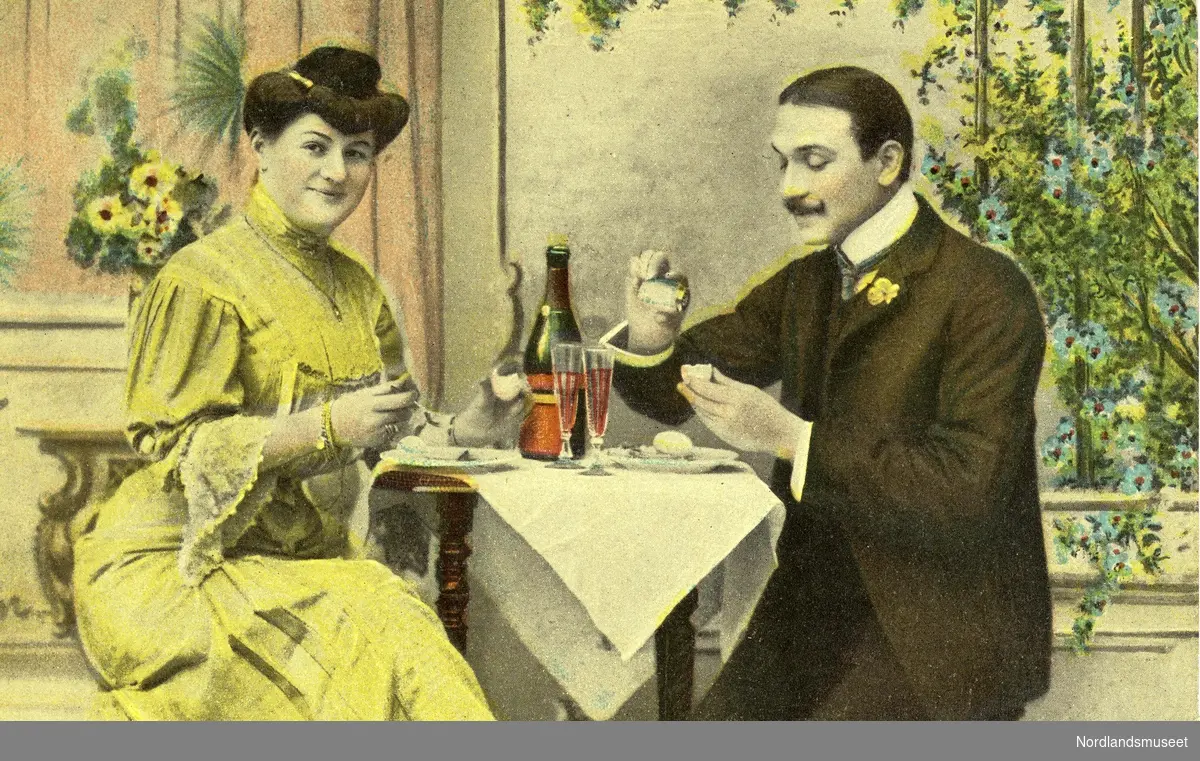 Postkortbilde. En mann og en kvinne som sitter ved et bord. Champagneflaske og to champagneglass på bordet. Hvit duk. Kvinnen har gul kjole og oppsatt hår, mannen har sort dres, hvit skjorte og gul knapphullspynt og en stor bart. I bakgrunnen er det lyserøde gardiner med gule blomster, og et vindu med planter.

Bakside: Adresse Frk. Kirsten Hansen, Grøtavær. "Carte postale" i rød skrift. Påskrevet dato 11.11.1907.
