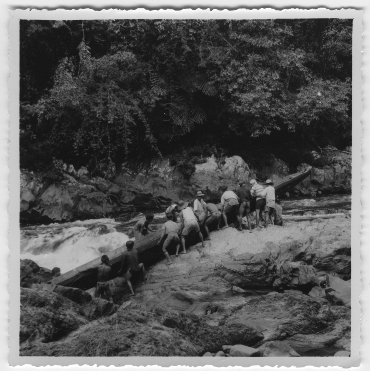 '14 st personer bär 1 kanot i floden. Text på baksidan: ''Vid ett vattenfall i Sembakongfloden. Kanoten måste dragas över land. R. Blomberg'' :: Ingår i serie med fotonr. 4038:1-4.'