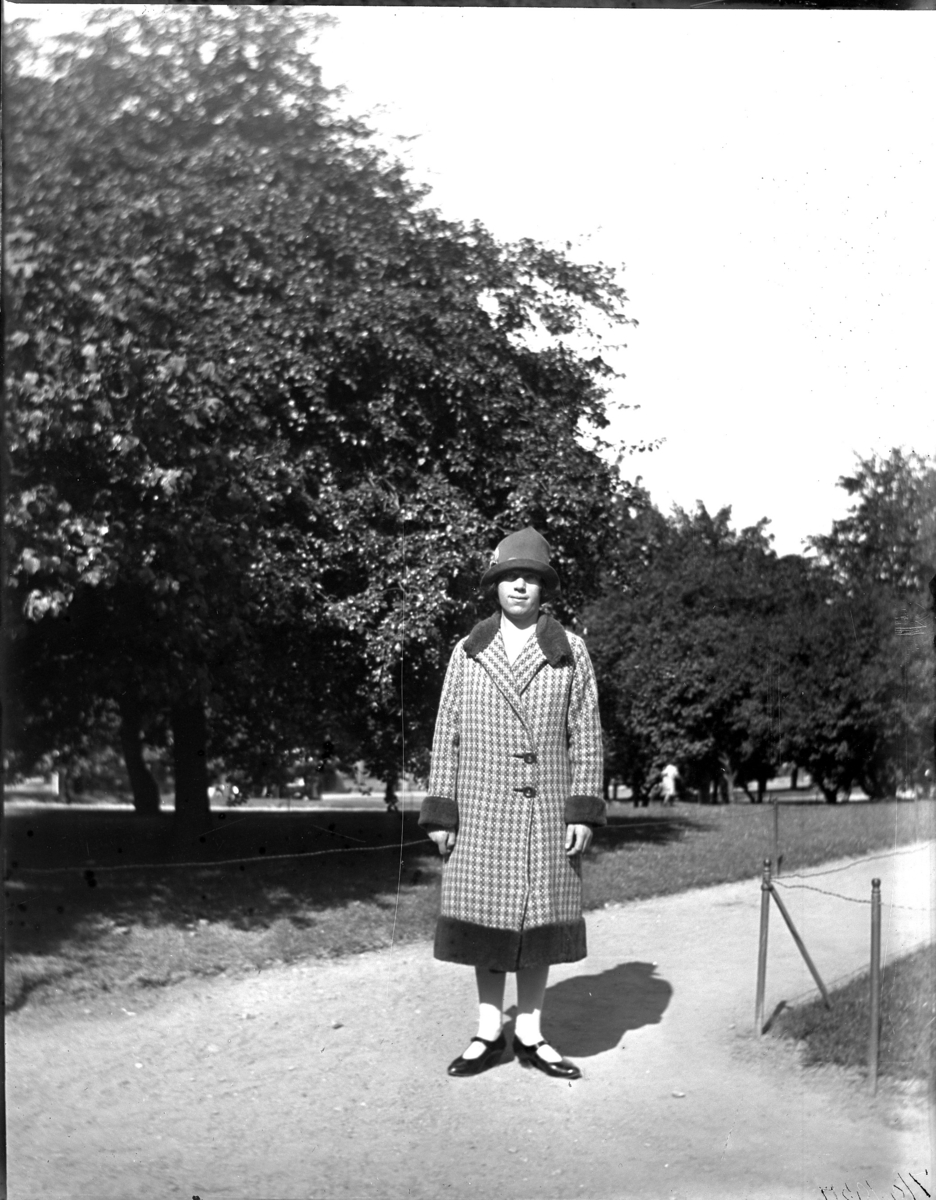 'Bildtext: ''Signe 7/9 1927.'' :: I kvinna iklädd kappa och hatt stående på grusgång. Vy med träd i bakgrunden. I en park. Klädsel. ::  :: Ingår i serie med fotonr. 5228:1-11.'