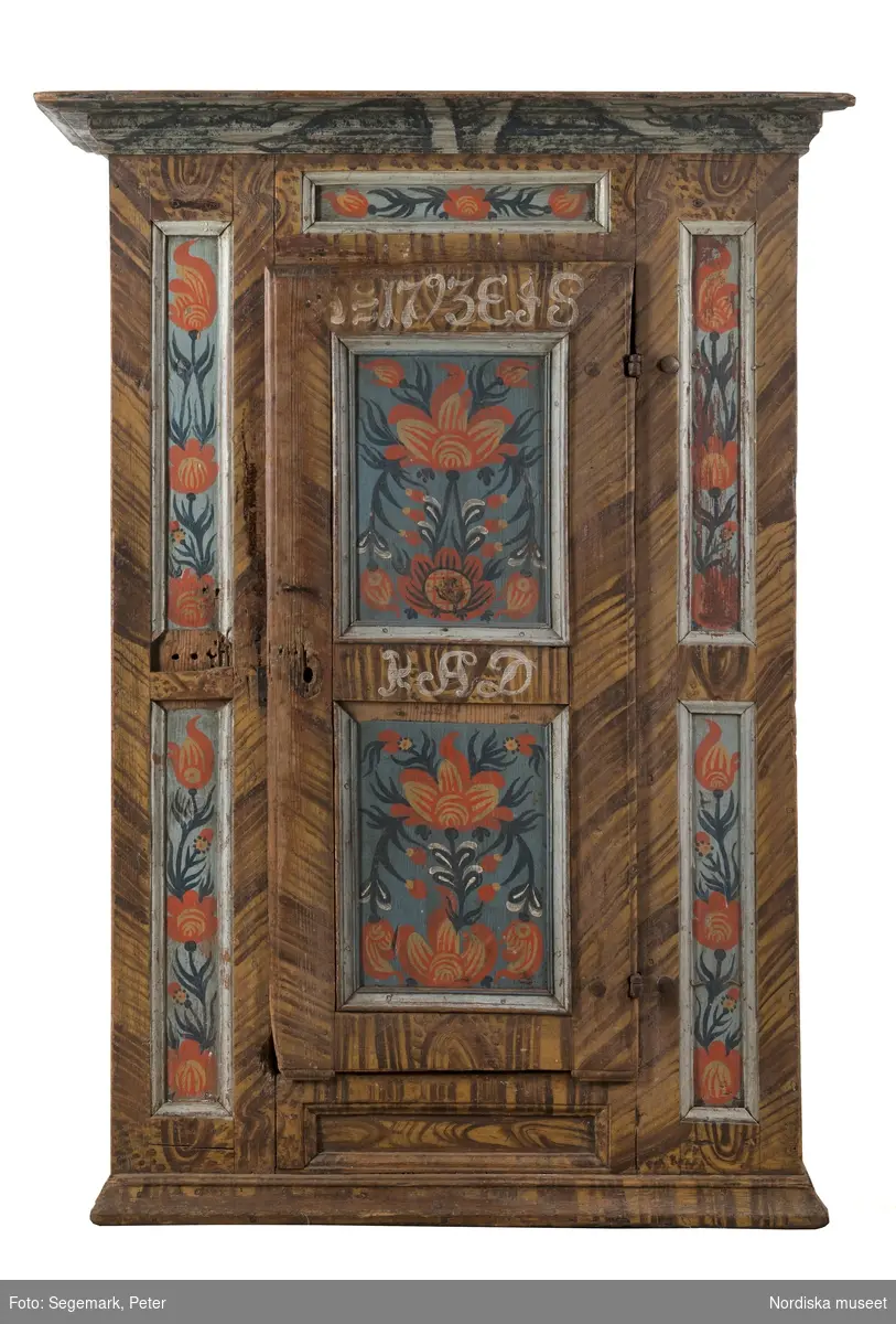 Katalogkort:
"Skåp av furu, med 1 dörr och 4 hyllor, brunmålat, med blommor å fälten, märkt Ao 1793 E. I. S. / K. A. D"