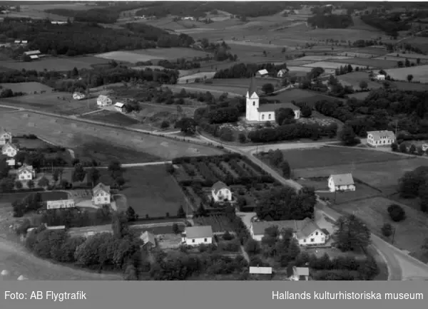 Flygbild över Valinge. Bybebyggelse med enfamiljshus och trädgårdar. Valinge kyrka och kyrkogård. Byvägar. Skog och odlingslandskap.