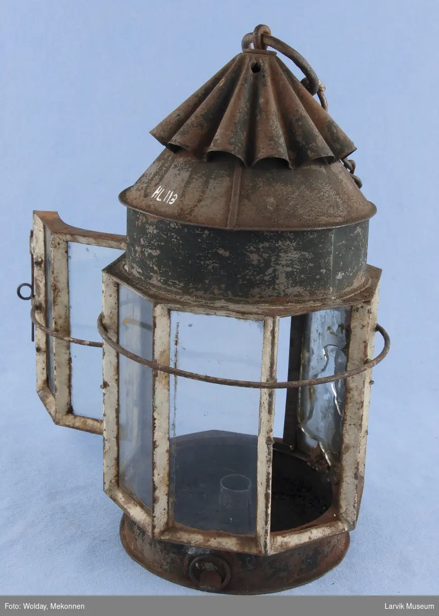 Sylinderformet lampe med bølgeformet toppstykke med ring for oppheng. 8 glassruter hvorav to utgjør dør.

