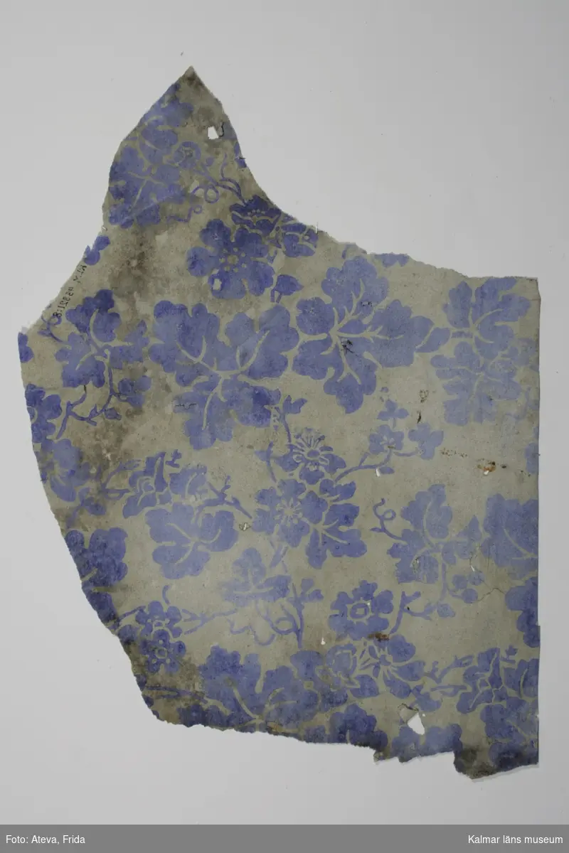 KLM 43821:8. Tapet av papper.Tryckt tapet med blommor och blad i ultramarinblått på ofärgad botten. På baksidan syns avtryck från schablonmålning i vinrött med större rutor. Även ljusgrå färg är det avtryck från. Datering: 1840-1850-tal.