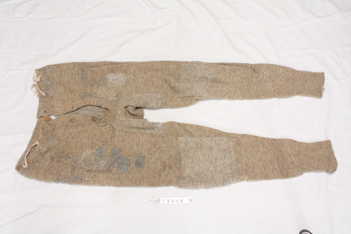 Lange strikkede ullunderbukser. Reparert, stoppet og lappet mange steder. To hemper på fremsiden trolig for å holde underbuksene på plass, enten med seler eller til buksen/genseren over.