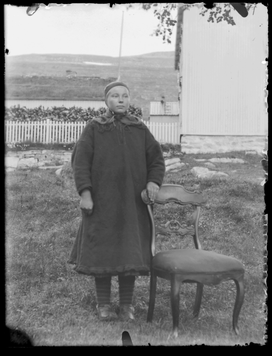 Samisk kvinne fotografert ute på tunet i kofte ved siden av en stol