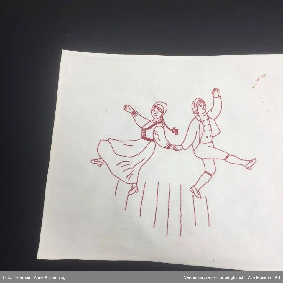 Rektangulært, hvit bordløper. Brodert motiv med rød tråd: En mann  som sitter og spiller trekskpill i midten, mellom to par som danser.