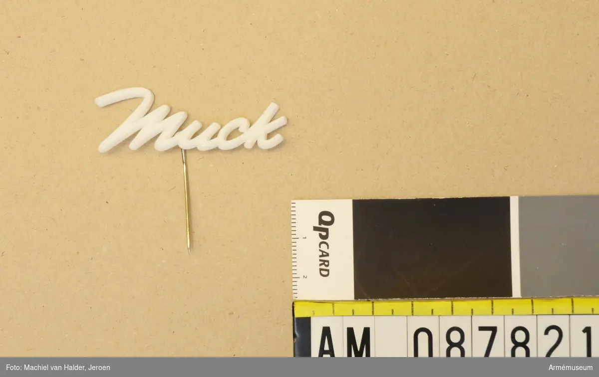 Vit pin i form av ordet "Muck", tillverkad i plast med nål i metall.