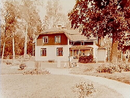 En och en halvvånings vinkelbyggt bostadshus med veranda.
Familj 5 personer och en hund framför huset.
Inspektor Theodor Egartz