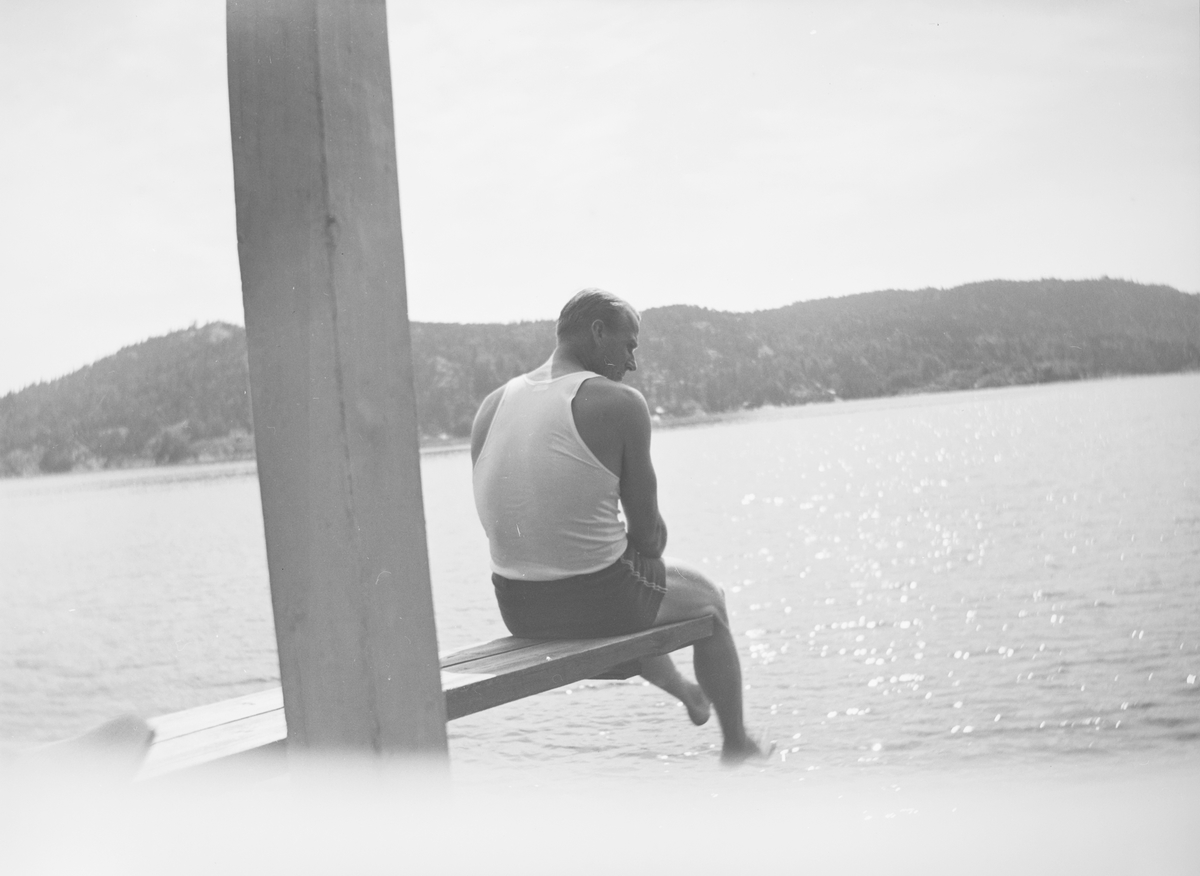 En stolpe deler bildet i to. En mann sitter ytterst på et stupebrett og ser ned mot vannet. Sola skinner.