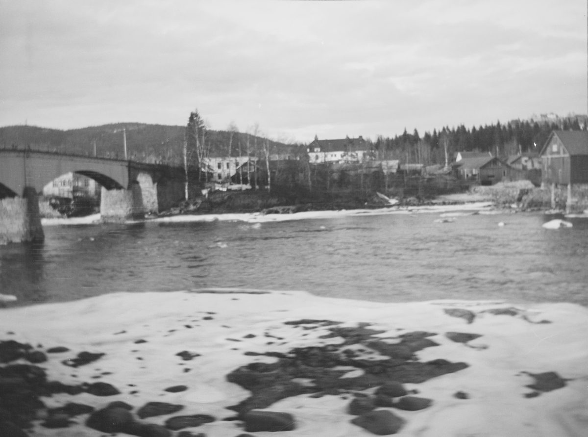 I front sees en snøkledt strand mot en elvestrøm med steiner. En bro går over elva. På den andre siden sees landlig bebyggelse. Hvite bjørkestammer lyser i landskapet.